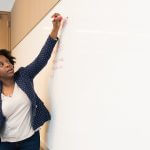 A woman holding a red marker and writing on the whiteboard.