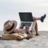 An image featuring a person wearing a hat while at the beach and using a laptop.