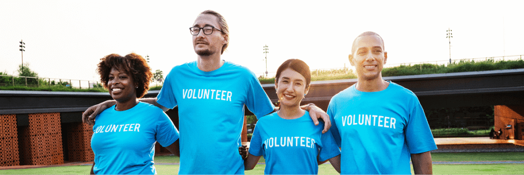 Group of volunteers in blue ‘VOLUNTEER’ shirts standing outdoors and smiling.