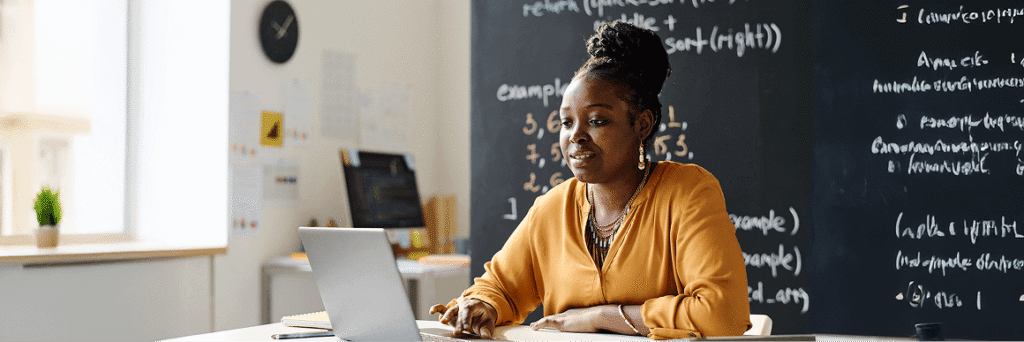 Educator working on a laptop in front of a chalkboard, creating or delivering training.