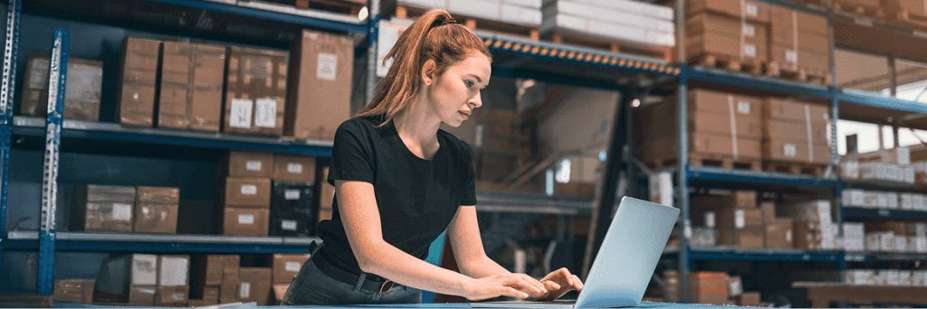 Person working on a laptop in a warehouse-like space with shelves behind them.