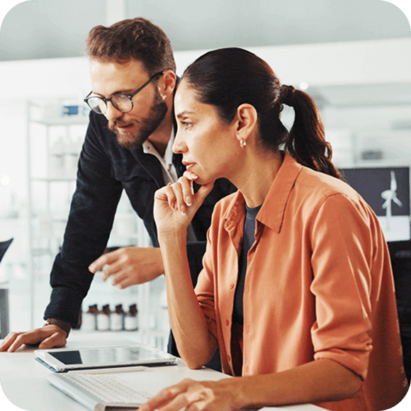 Two coworkers collaborating at a laptop, one standing and one seated, focused on the screen.