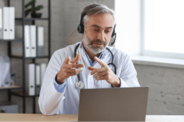 A male doctor wearing a headset and gesturing while speaking into his laptop during a remote video session.