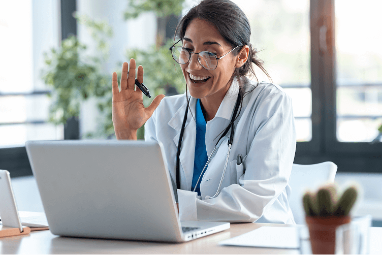 A female healthcare professional in a white lab coat smiling and waving at a laptop during a virtual training session.