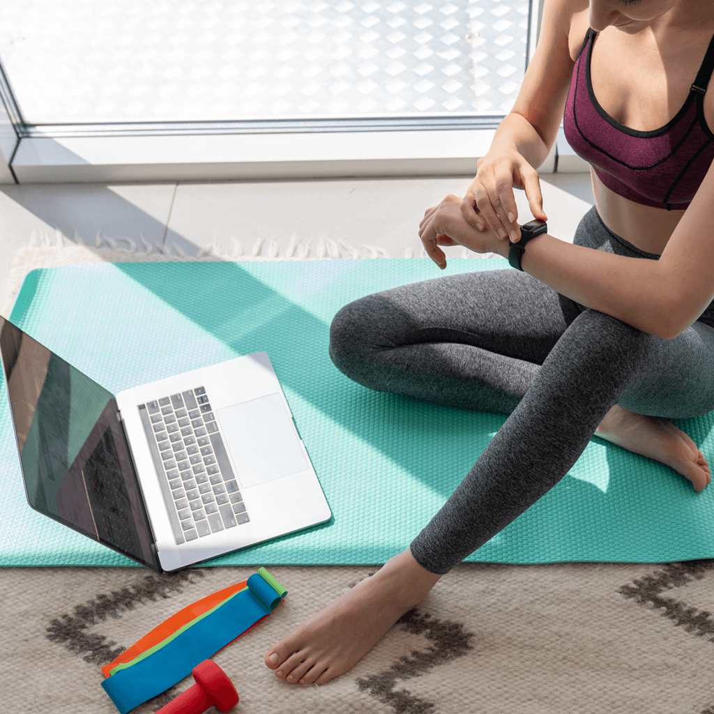 Person sitting on a yoga mat checking a smartwatch beside an open laptop, with resistance band and small dumbbells on the floor.